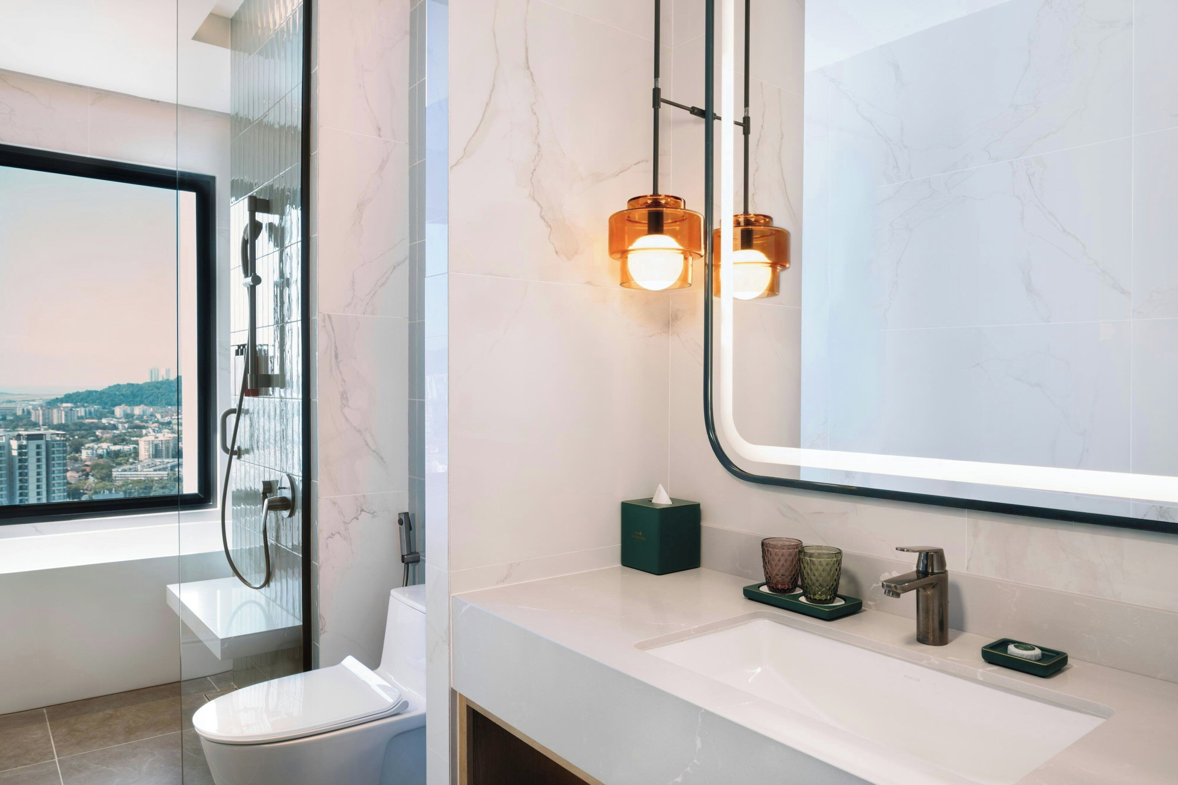 Een moderne badkamer met een douchecabine van glas, een toilet, een wastafel met spiegel en uitzicht op een stadsgezicht door een raam.