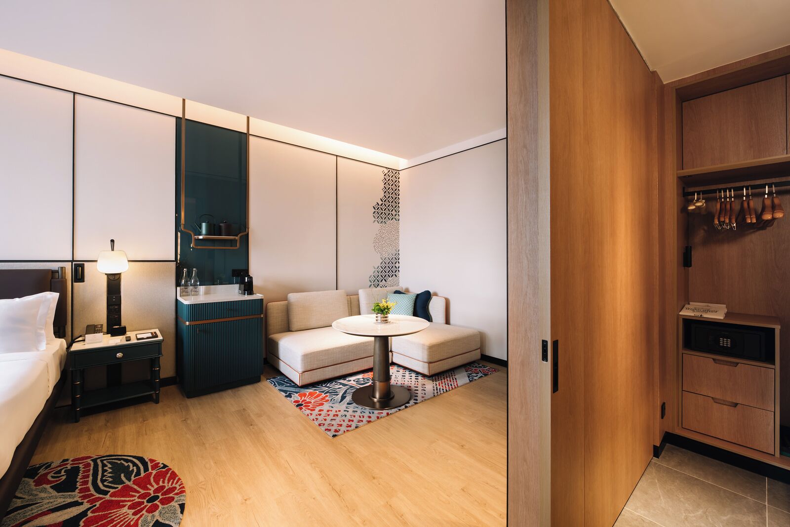 A modern hotel room with a bed, a sofa, a small table, a desk with a lamp, a minibar, and a wardrobe.