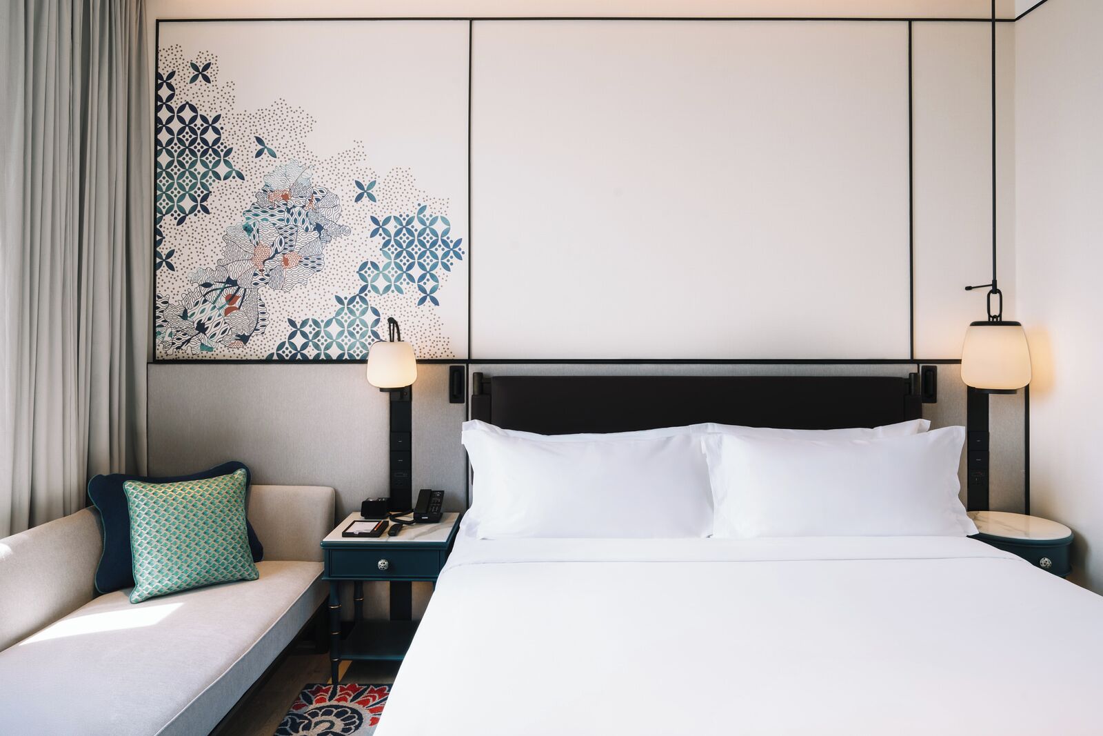 A hotel room with a bed, a nightstand, a lamp, a sofa with a pillow, and a wall art piece.