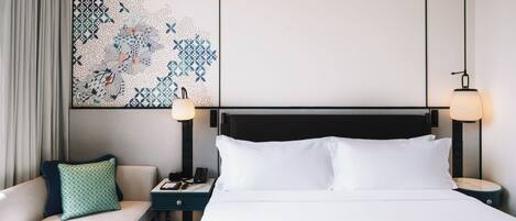 A hotel room with a bed, a nightstand, a lamp, a sofa with a pillow, and a wall art piece.