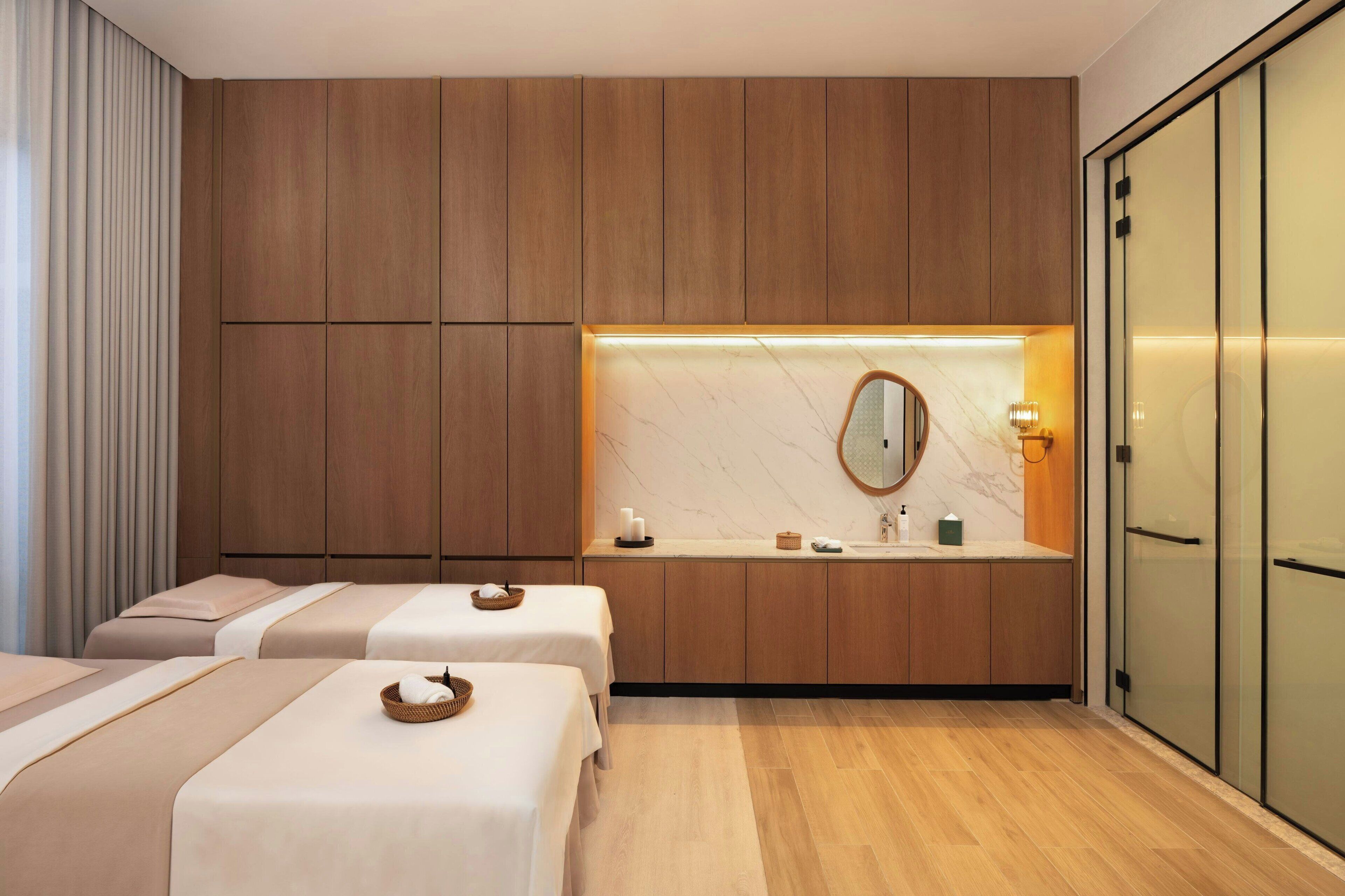 A hotel room with two beds, a wooden wardrobe, a marble-topped vanity, and a glass door closet.