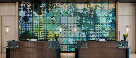 Zona de recepción de hotel con una gran pared de mosaicos, dos mostradores y luminarias modernas.