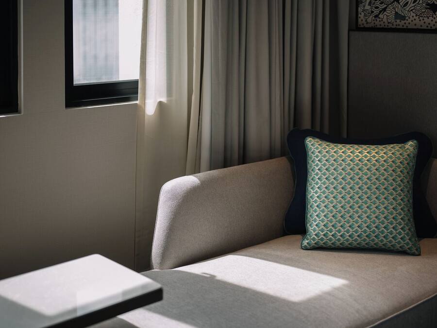 A modern hotel room with a sofa, a patterned pillow, and a window with curtains.