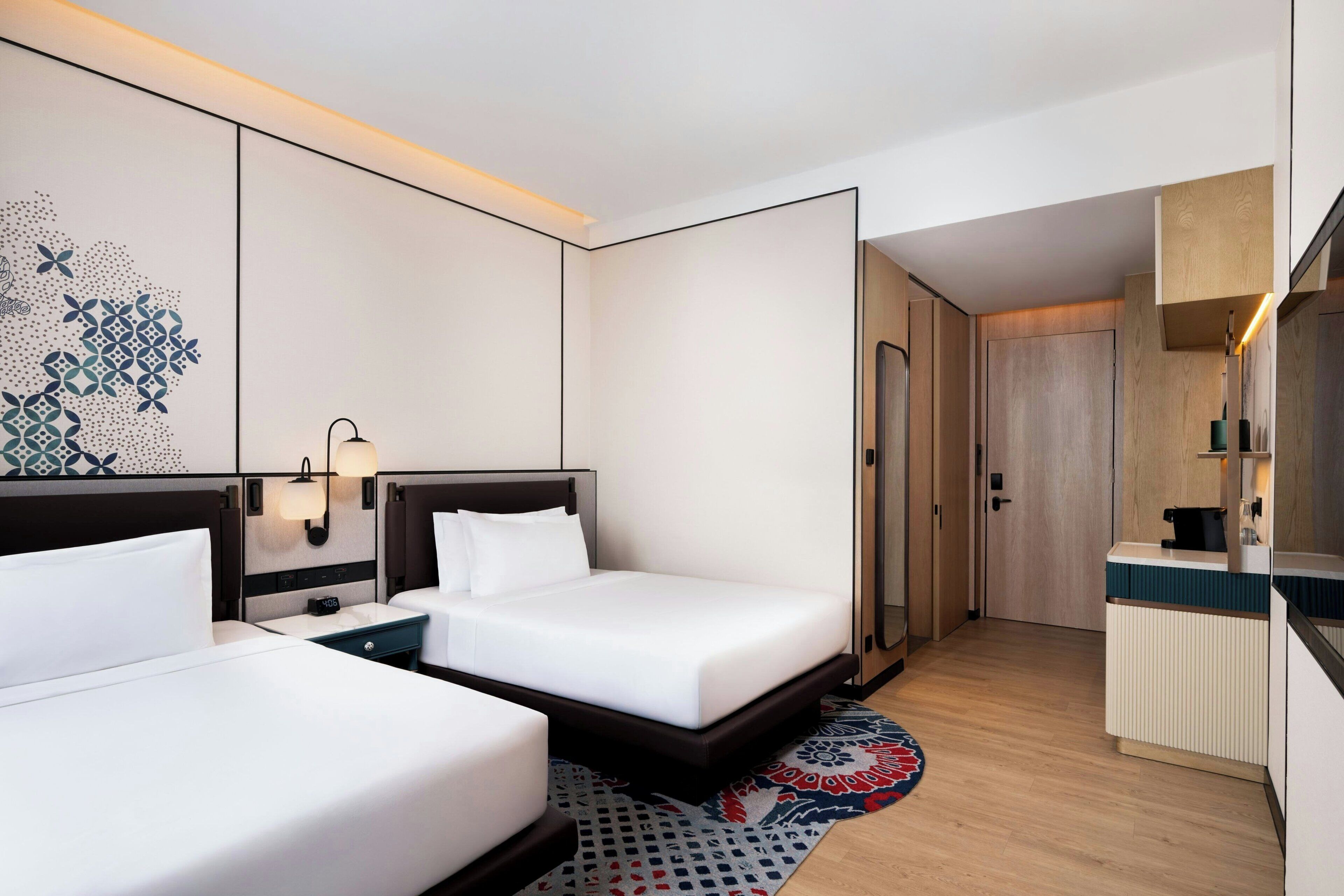 Una camera d'albergo con due letti, una scrivania, uno specchio e l'ingresso del bagno.