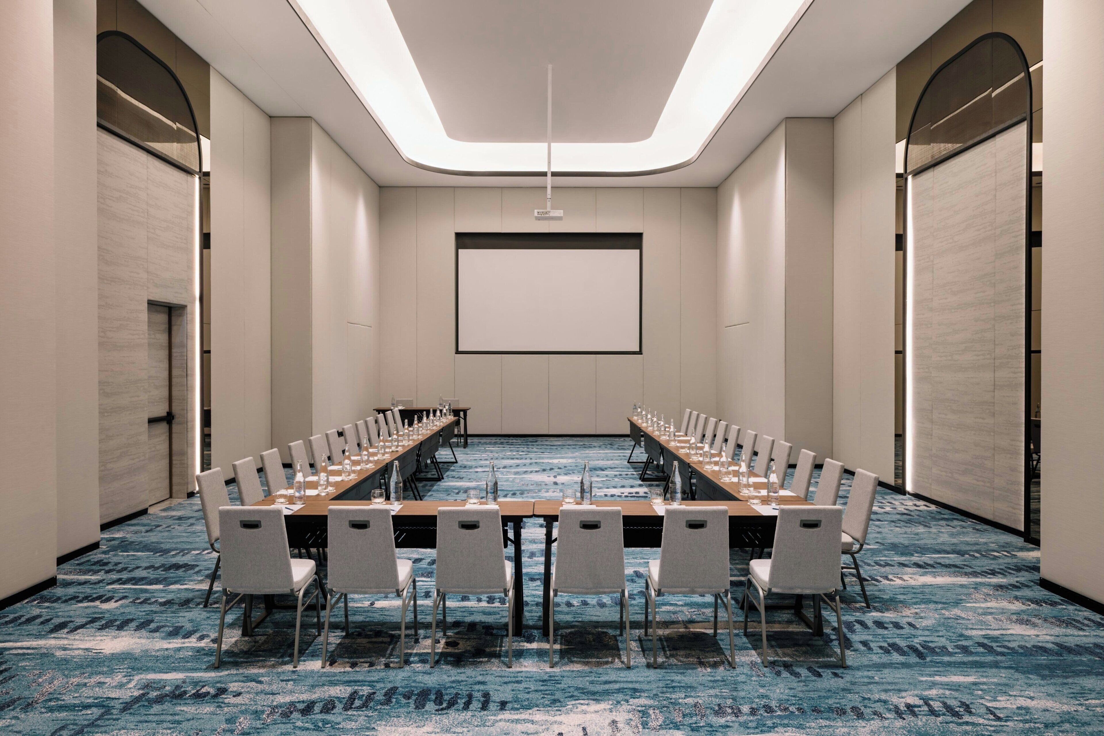 A conference room with a long table and chairs, a projector screen, and a carpeted floor.