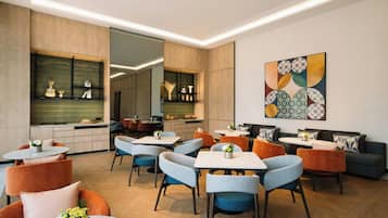 Uma área de refeições moderna com várias mesas e cadeiras, uma cozinha ao fundo e um elemento decorativo na parede.