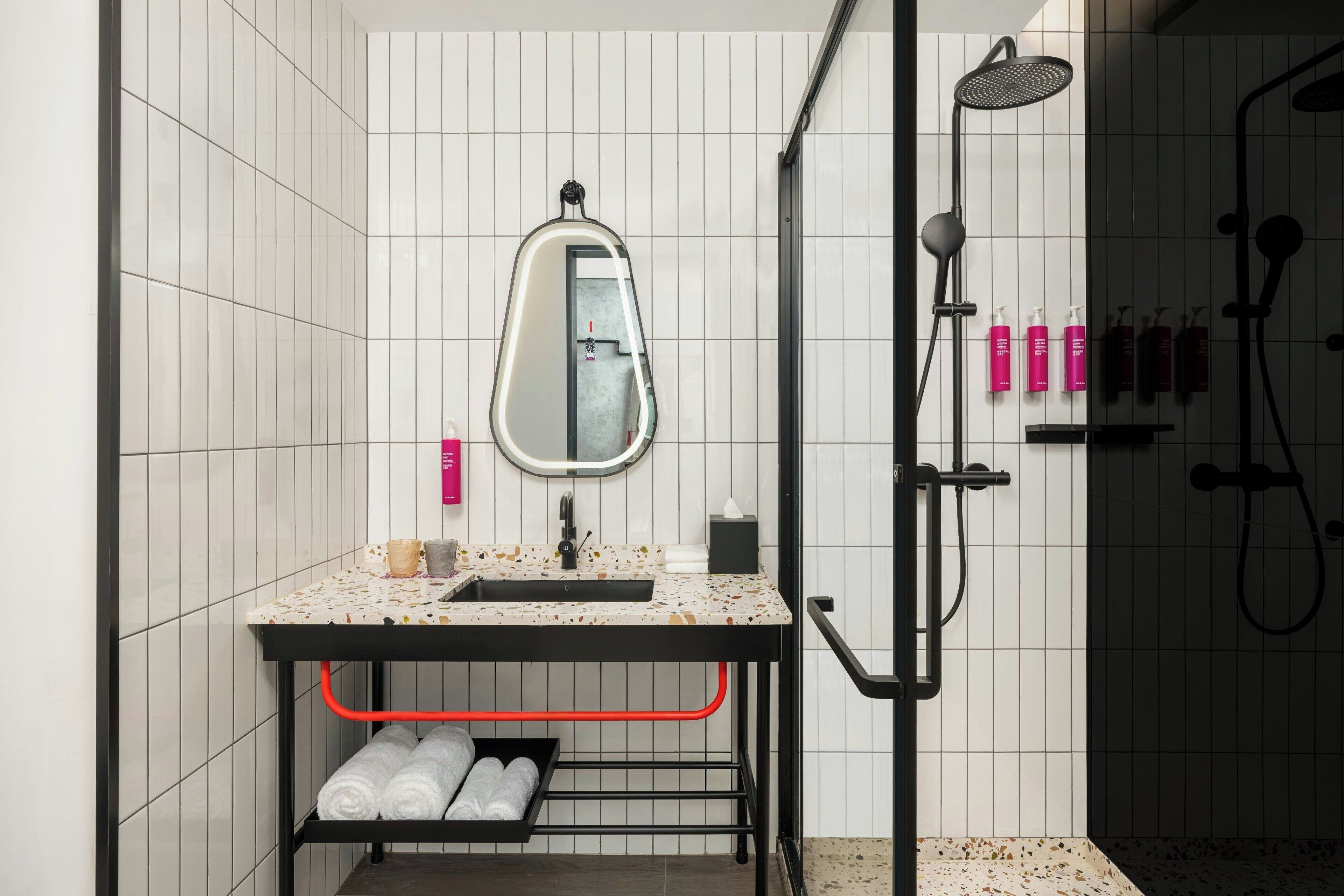 Ein modernes Badezimmer mit weiß gefliester Wand, einem Waschbecken mit Spiegel, einer Duschkabine und ordentlich gerollten Handtüchern auf einem Regal.