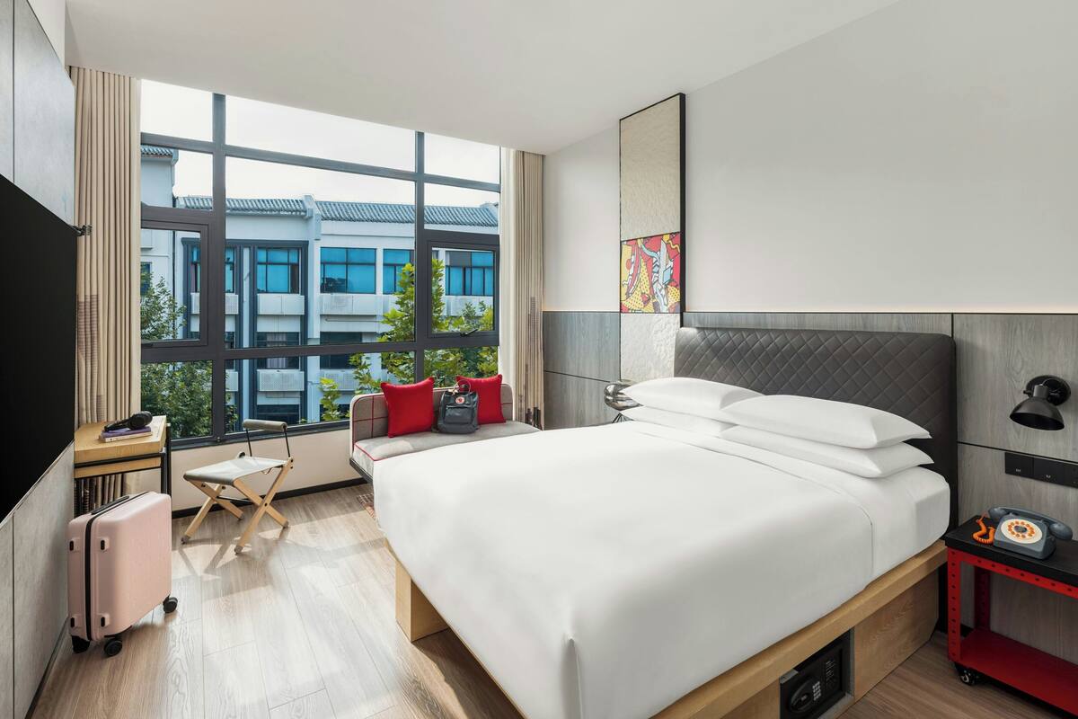 A modern hotel room with a large bed, a desk with a chair, a suitcase, and a view of buildings outside.
