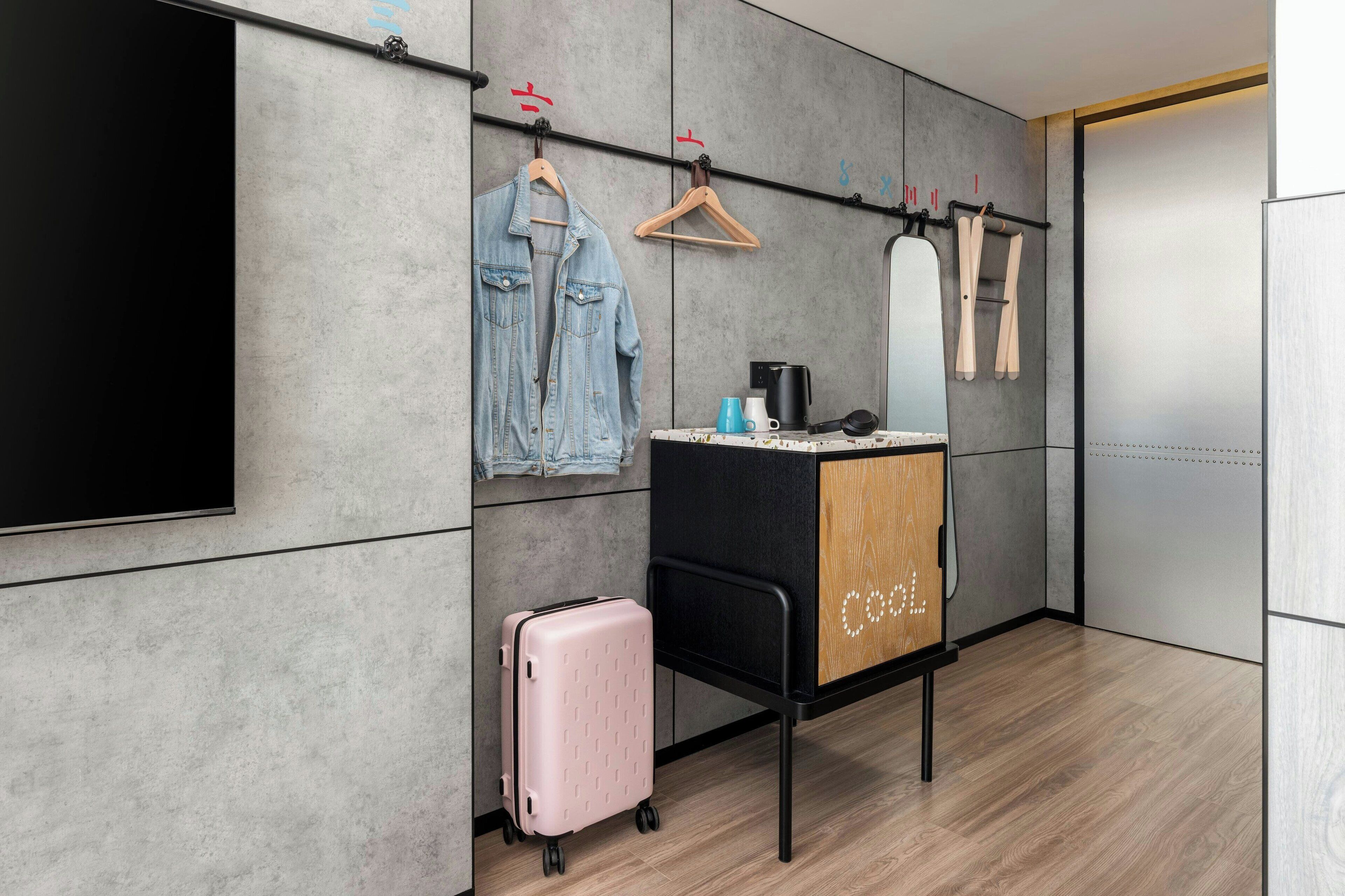 A modern hotel room with a flat-screen TV, a wooden cabinet with a mirror, a hanging rack with clothes, a suitcase, and a coffee machine.