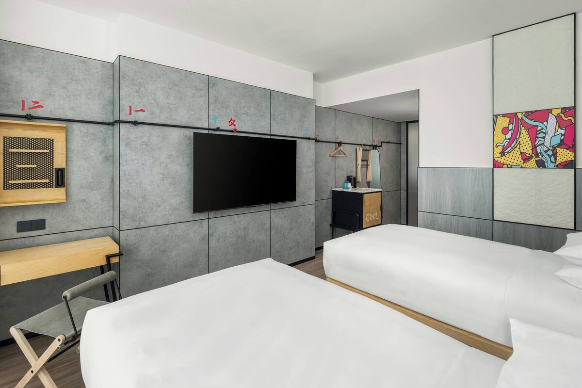 A modern hotel room with two beds, a flat-screen TV mounted on the wall, a desk with a chair, and a small seating area.