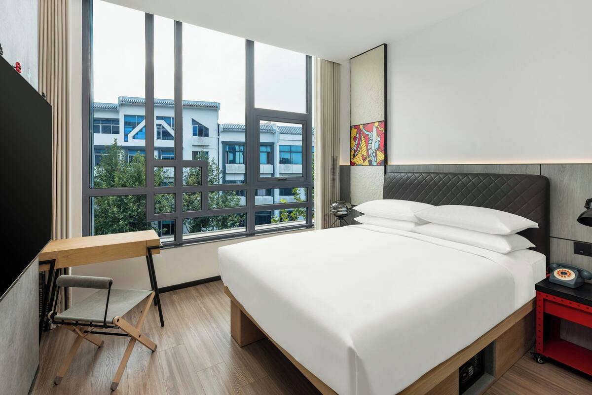 A modern hotel room with a large window, a bed with white linens, a desk with a chair, and a red nightstand.