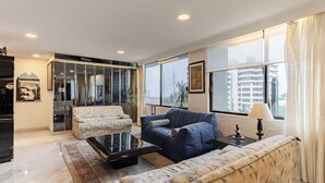 Un saló modern amb un sofà, una butaca i una taula de centre. Hi ha un finestral gran amb cortines, una porta que condueix a una altra habitació i una taula petita amb una làmpada.
