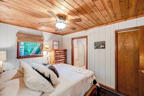 Camera da letto con rivestimenti in legno, un letto, una ventola a soffitto, una finestra con persiane e un comò in legno.