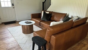 Un sofá de cuero marrón con un cojín blanco y negro, una mesa de centro redonda de madera y un taburete negro.