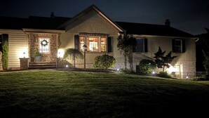 Una casa de dos pisos con fachada de piedra, un porche con un banco y una puerta principal iluminada.