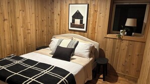 Un dormitorio con cama, mesita de noche, lámpara junto a la ventana y un cuadro en la pared.
