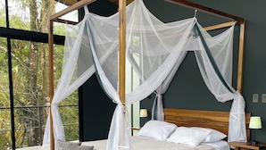 Un lit à baldaquin à quatre poteaux, doté d’une structure en bois et d’un ciel de lit blanc, un tapis vert et une vue sur la verdure à travers de grandes fenêtres.