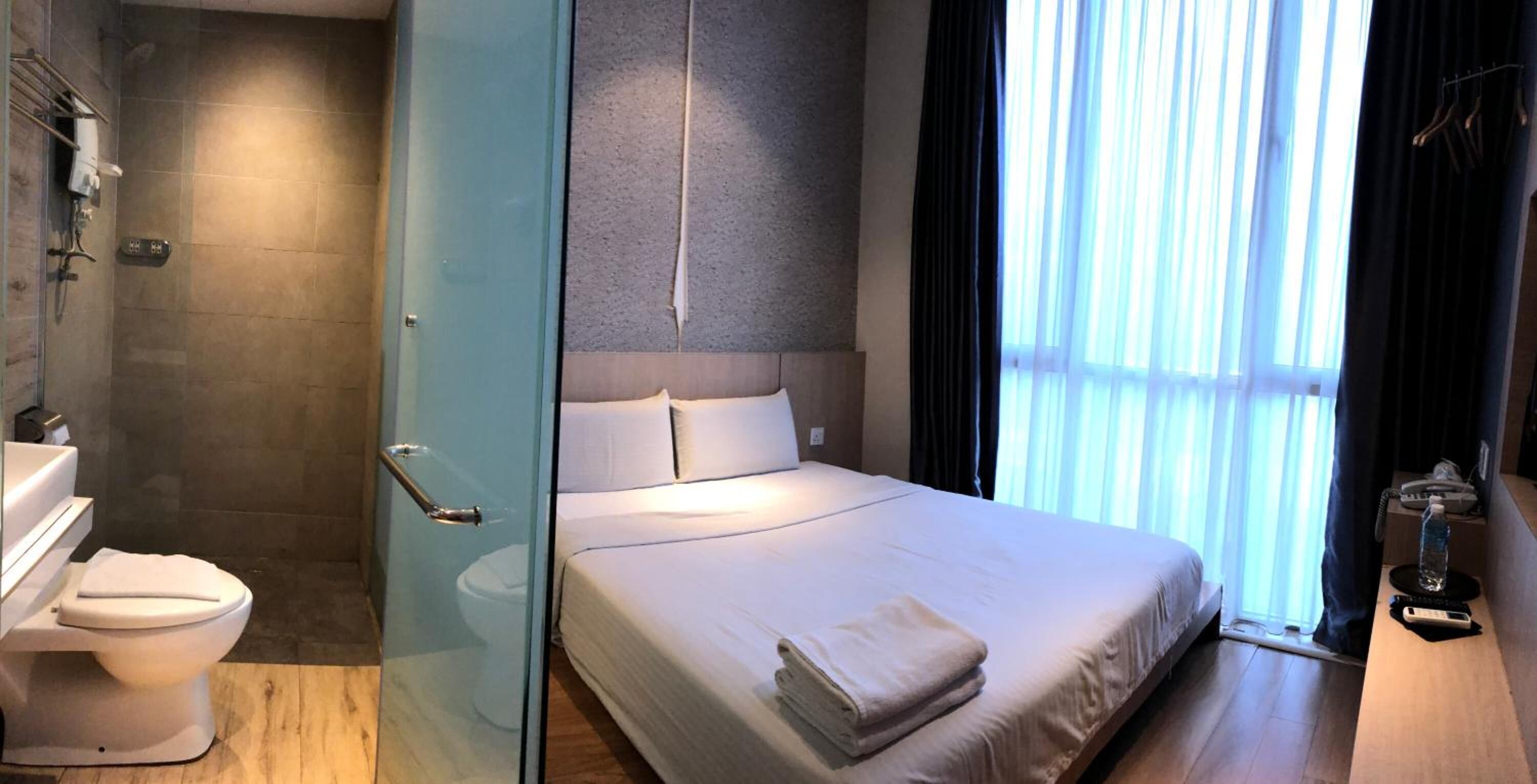 Habitación de hotel moderna con separador de vidrio que divide el baño y el dormitorio. La habitación cuenta con una cama grande, una mesita de noche y vistas al exterior a través de cortinas translúcidas.