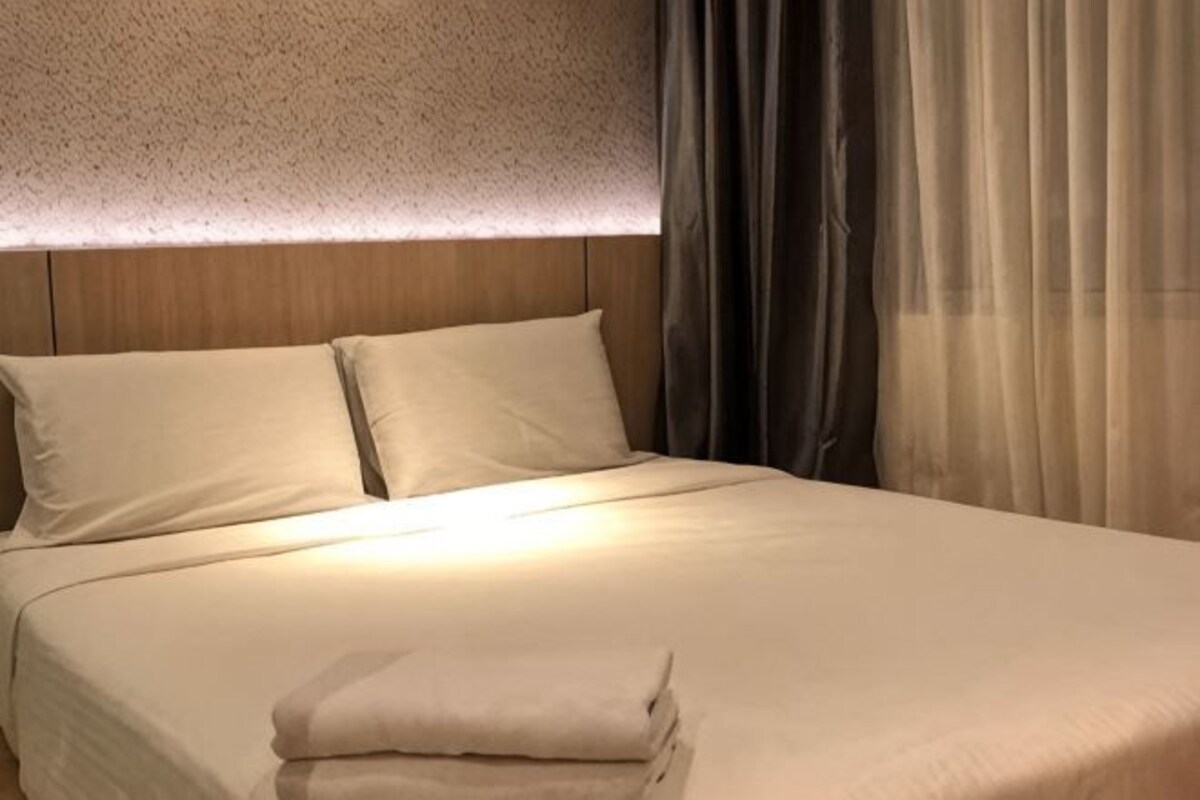 Cama bem arrumada com almofadas e um cobertor dobrado, cabeceira de madeira e cortinas leves.