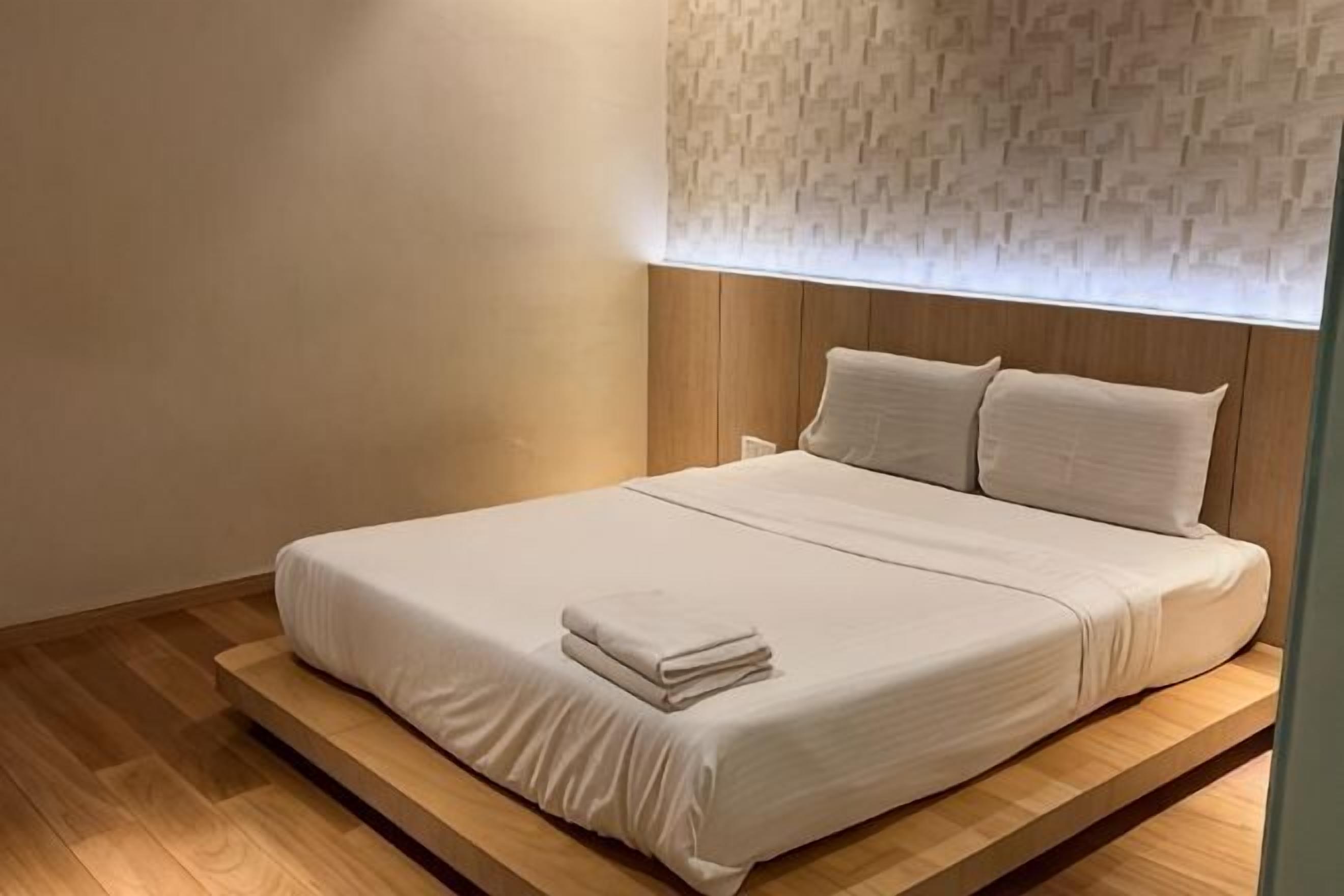 Una cama bien hecha con una colcha blanca y almohadas, una plataforma de madera debajo y un papel tapiz estampado en la pared.