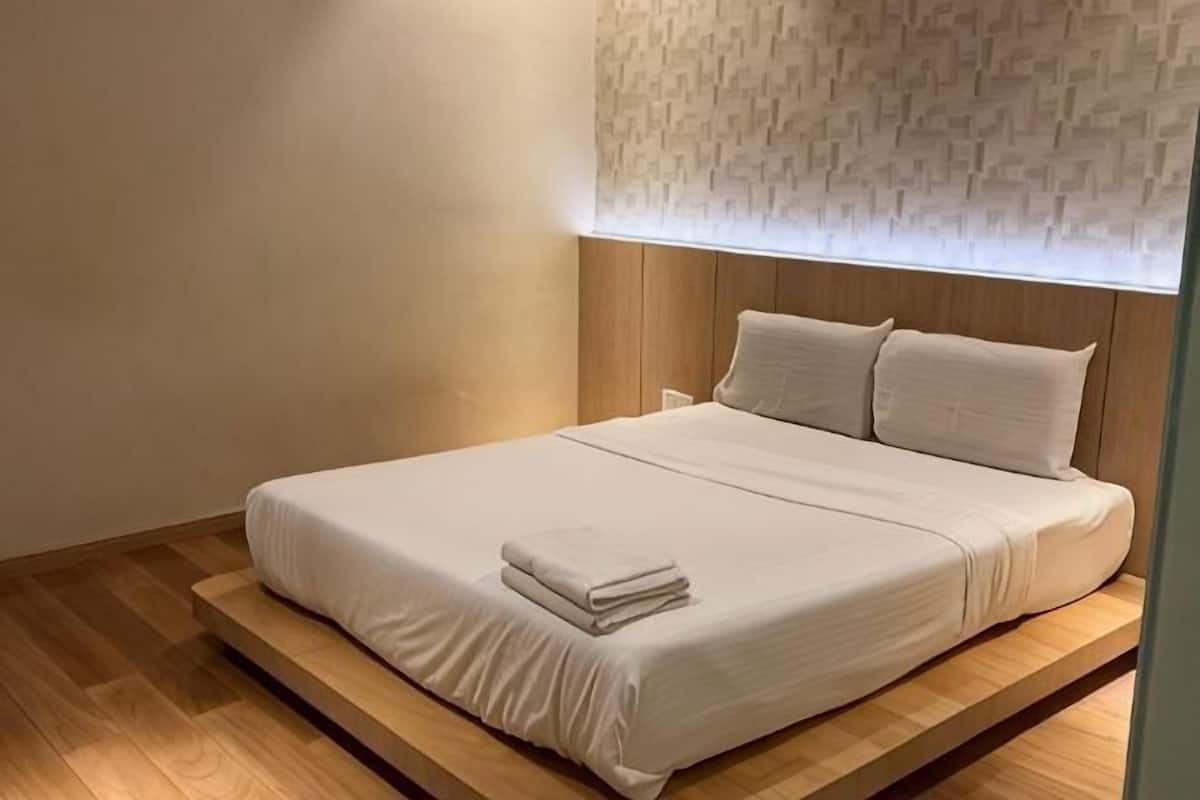 A neatly made bed with a white bedspread and pillows, a wooden platform under the bed, and a patterned wall above.