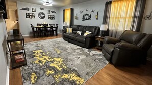 A living room with a black leather sofa, a grey and yellow rug, a dining area with a table and chairs, and wall decorations including clocks and framed pictures. - Cute 3 bedroom, 1 bath home less than 5 minutes from downtown. (Hot Springs)