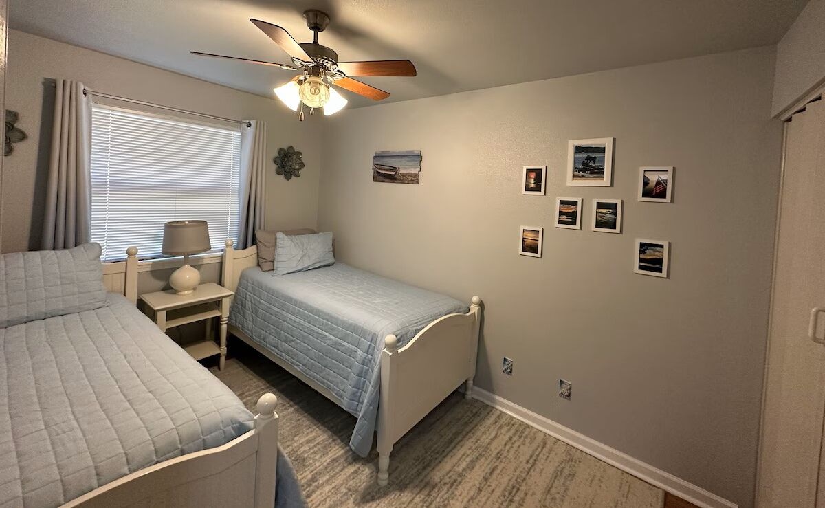 Una camera da letto con due letti, una ventola a soffitto, una finestra con tende e diverse foto incorniciate appese al muro.