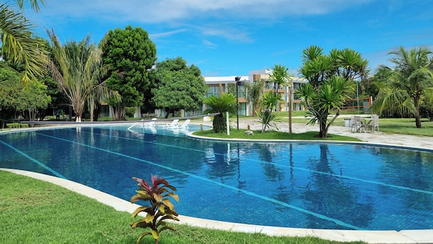 A swimming pool with clear blue water, surrounded by lush green grass and palm trees. - Suíte + Quarto com Ar, Wi-Fi e 2 Vagas (Barra de São Miguel)