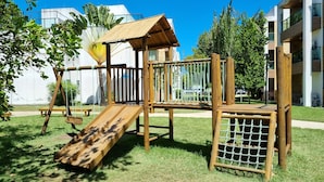 A wooden playground with a slide and climbing net, surrounded by grass and trees, with modern buildings in the background. - Suíte + Quarto com Ar, Wi-Fi e 2 Vagas (Barra de São Miguel)