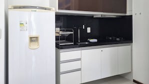 A compact kitchen with a white refrigerator, a black countertop, and a stainless steel sink. - Suíte + Quarto com Ar, Wi-Fi e 2 Vagas (Barra de São Miguel)