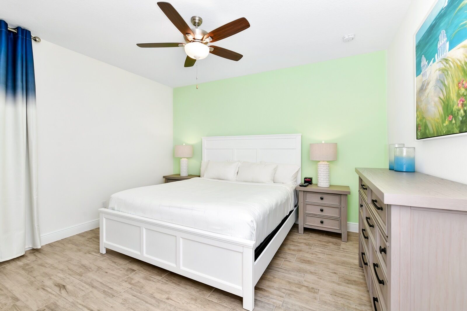 A bedroom with a bed, nightstands, a dresser, a ceiling fan, and a painting on the wall.