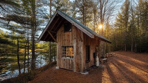 Eine rustikale Holzhütte mit geneigtem Dach, einem Fenster und einer Tür, umgeben von einem Wald und einem Gewässer.