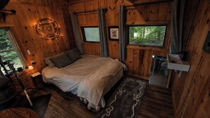 Ein Schlafzimmer einer Holzhütte mit einem Bett, einem kleinen Tisch, einem Waschbecken und einem Fenster mit Blick auf Grünflächen.