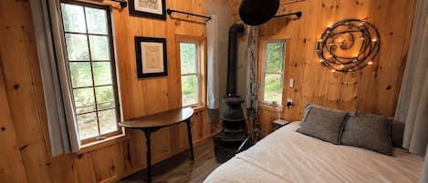 A wooden cabin bedroom with a bed, a table, a stove, and framed pictures on the wall.