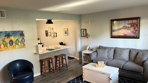 Un salón con un sofá gris, una silla azul, taburetes de madera, una mesa de centro blanca con dados y un cuadro enmarcado de Chicago.