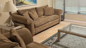 A living room with a beige couch, a glass coffee table, a lamp, and a painting of a beach scene. - Chic 2-bedroom condo with AC, WiFi in Naples, Resort like pool. 3 miles to beach (Naples)