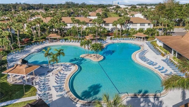A large, kidney-shaped swimming pool with multiple sunbathing areas and palm trees, surrounded by resort buildings. - Chic 2-bedroom condo with AC, WiFi in Naples, Resort like pool. 3 miles to beach (Naples)