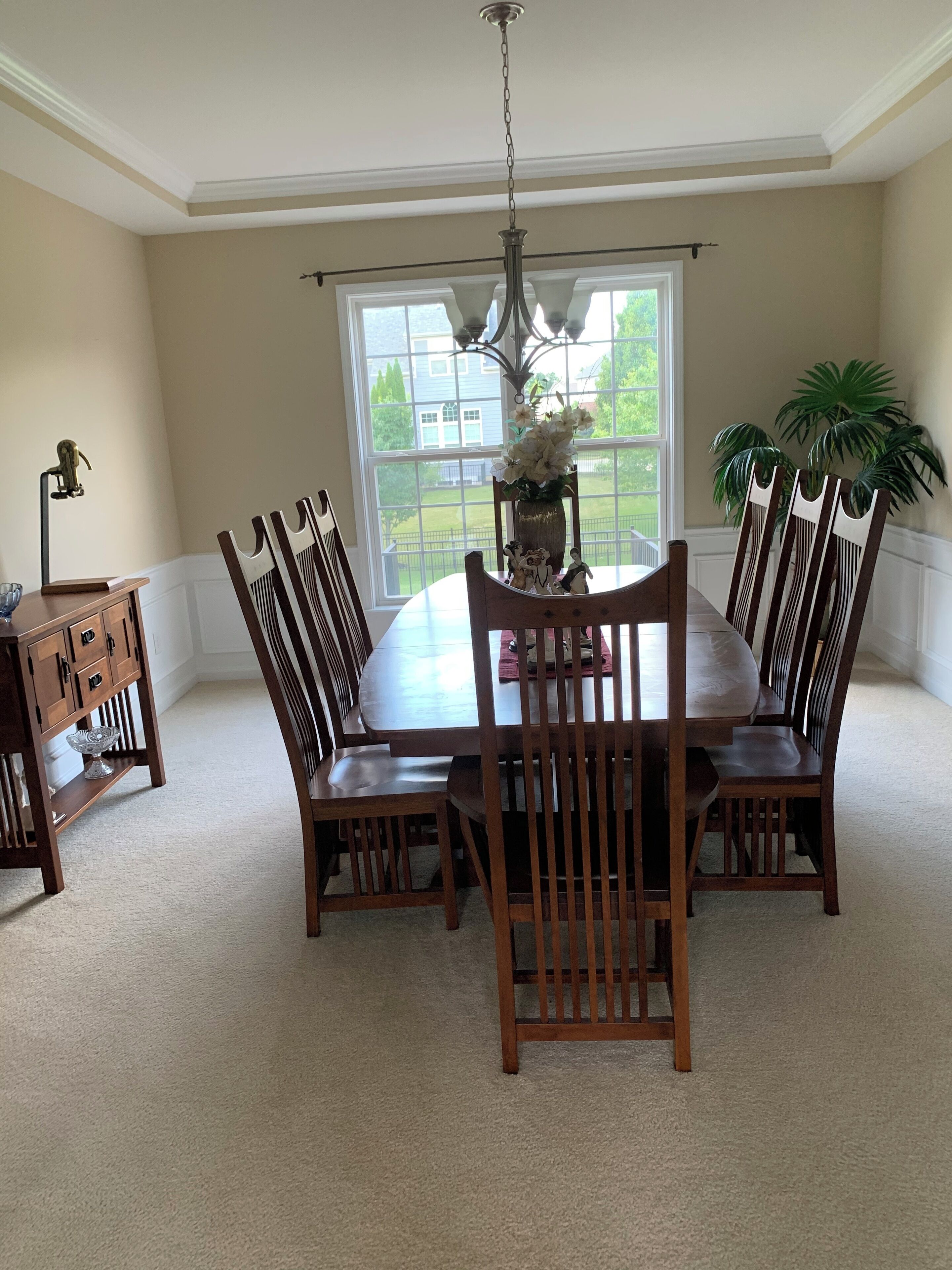 A dining room with a large table, six chairs, a chandelier, a vase with flowers, and a potted plant.