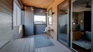 Un espace extérieur abrité doté d’une terrasse en bois, d’un petit coin salon avec une chaise et une table, et d’une porte-fenêtre coulissante donnant sur un espace intérieur équipé d’un ventilateur de plafond.