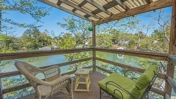 A covered outdoor seating area with wicker chairs and a green cushioned bench, overlooking a body of water and greenery. - Charming studio in lovely Topping with AC, WiFi that has stunning water views! (Topping)