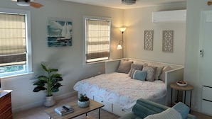 A bedroom with a bed, bedside tables, a ceiling fan, a window with blinds, a wall-mounted air conditioner, and a painting. - Charming studio in lovely Topping with AC, WiFi that has stunning water views! (Topping)