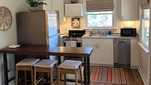 A modern kitchen with white cabinets, a stainless steel refrigerator, a microwave, and a dishwasher. There is a wooden dining table with two stools and a ceiling fan. - Charming studio in lovely Topping with AC, WiFi that has stunning water views! (Topping)