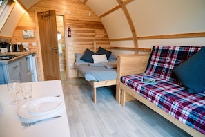 Un intérieur compact en bois, comprenant un coin cuisine, un lit avec une couette à carreaux et un coin repas avec une table blanche dressée.