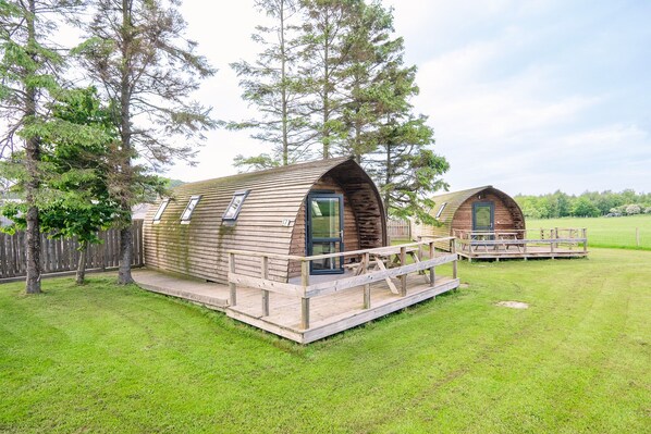 Two wooden cabins with a deck, surrounded by trees and a grassy field. - Cosy Rural Cabin With Accessible Facilities (Kinloss)
