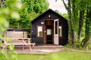 A small, dark wooden cabin with a number 13 sign, a wooden bench, and a hot tub in the foreground.