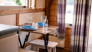 A wooden table with a blue plate, two glasses, and salt and pepper shakers in a room with wooden walls and a window. - Rustic Log Cabin With Hot Tub (Kinloss)