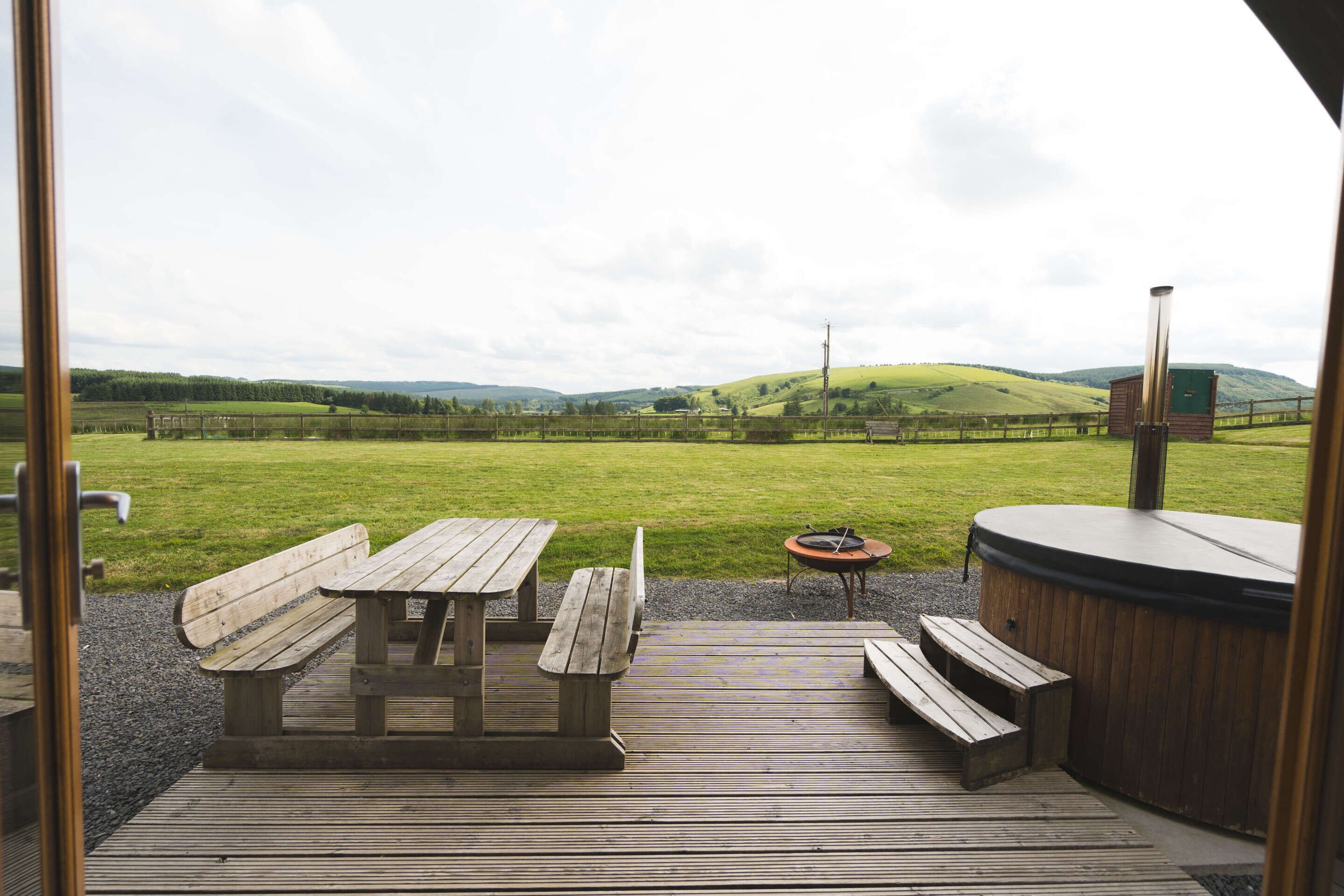 A wooden deck with picnic tables and a hot tub, overlooking a grassy field and hills.