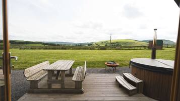 A wooden deck with picnic tables and a hot tub, overlooking a grassy field and hills.