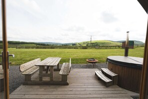 A wooden deck with picnic tables and a hot tub, overlooking a grassy field and hills. - Cosy Cabin with Hot Tub in the Welsh Countryside (Llanbrynmair)