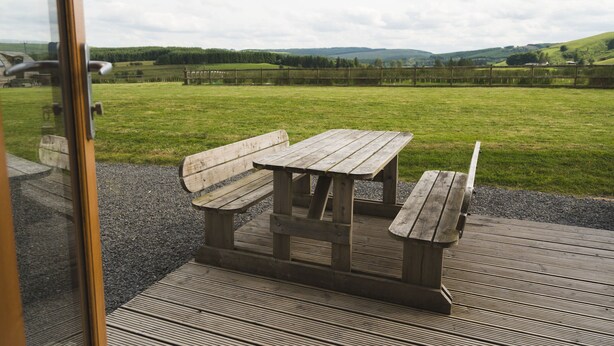 A wooden picnic table with benches on a wooden deck, overlooking a grassy field with a fence and distant hills. - Rural Cabin in the Welsh Countryside (Llanbrynmair)